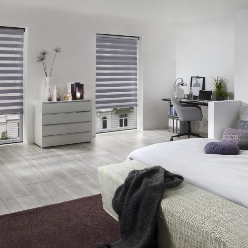 Modernes Schlafzimmer mit grauem Doppelrollo in der Stadt, Bett mit Kissen und Decke, Teppich, Kommode mit Deko und Schreibtisch in der Ecke